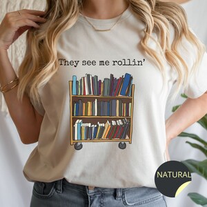 May include: A white t-shirt featuring the text "They see me rollin'" and a graphic of a rolling bookshelf filled with books. The t-shirt is available in the colour "Natural".