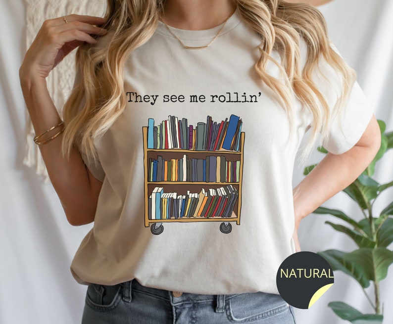 May include: A white t-shirt featuring the text "They see me rollin'" and a graphic of a rolling bookshelf filled with books. The t-shirt is available in the colour "Natural".