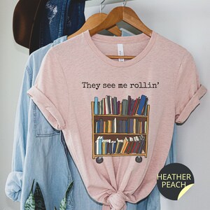 May include: A pink t-shirt with the text "They see me rollin'" and a graphic of a bookshelf on wheels filled with books. The text "HEATHER PEACH" is on a black circle sticker on the bottom right corner of the shirt.