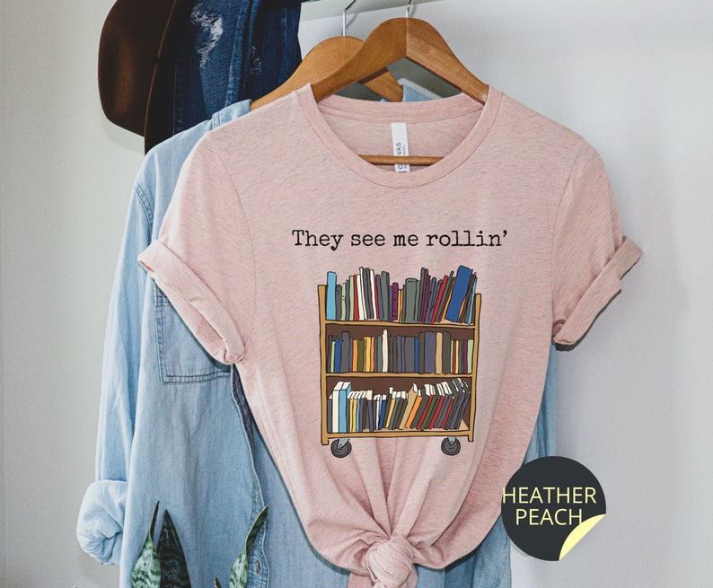 May include: A pink t-shirt with the text "They see me rollin'" and a graphic of a bookshelf on wheels filled with books. The text "HEATHER PEACH" is on a black circle sticker on the bottom right corner of the shirt.