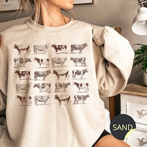 May include: A beige sweatshirt with a graphic print of 20 brown and white cows. The sweatshirt has a crew neck and long sleeves. The word "SAND" is visible on the bottom right corner of the image.