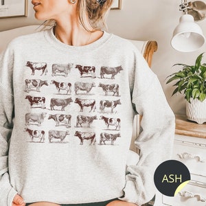 May include: A gray sweatshirt with a vintage-style print of cows in various poses. The print is in shades of brown and black, and the sweatshirt has a relaxed fit.