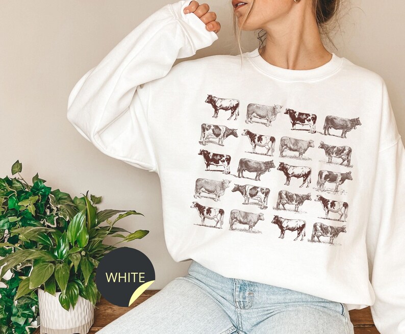 May include: White long-sleeved sweatshirt with a brown graphic print of multiple cows. The sweatshirt has a crew neck and is made of a soft, comfortable fabric.