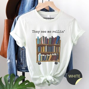 May include: A white t-shirt featuring the text "They see me rollin'" and a cartoon illustration of a rolling bookshelf filled with books. The t-shirt is on a hanger with a denim jacket and a brown leather bag.