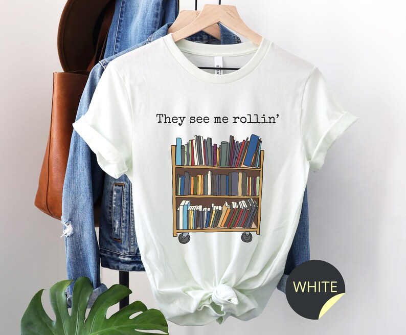 May include: A white t-shirt featuring the text "They see me rollin'" and a cartoon illustration of a rolling bookshelf filled with books. The t-shirt is on a hanger with a denim jacket and a brown leather bag.