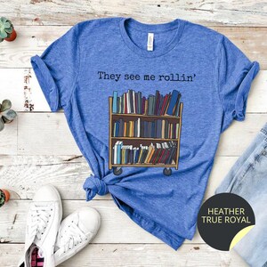 May include: A blue t-shirt featuring the text "They see me rollin'" and a cartoon image of a book trolley full of books. The book trolley has wheels and is on a wooden floor. The t-shirt is on a wooden surface with a pair of white trainers and a pair of blue jeans.
