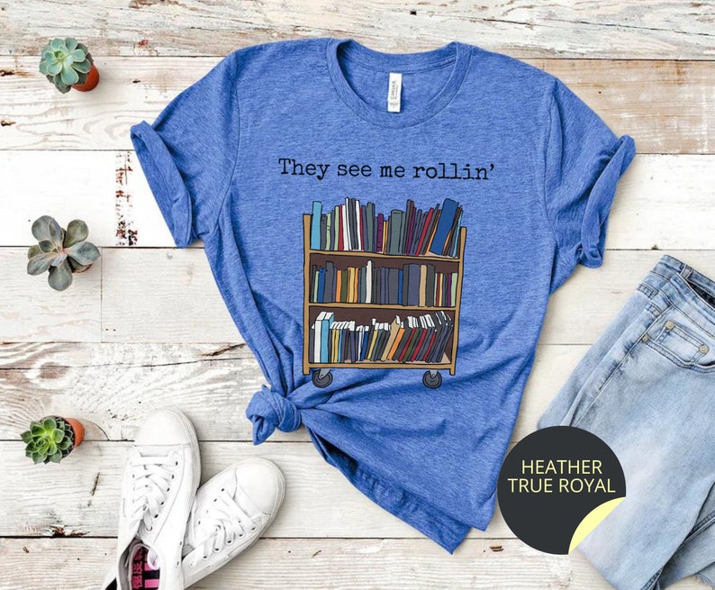 May include: A blue t-shirt featuring the text "They see me rollin'" and a cartoon image of a book trolley full of books. The book trolley has wheels and is on a wooden floor. The t-shirt is on a wooden surface with a pair of white trainers and a pair of blue jeans.