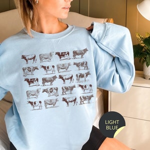 May include: A light blue sweatshirt with a pattern of brown cows printed on the front. The sweatshirt has a crew neck and long sleeves.