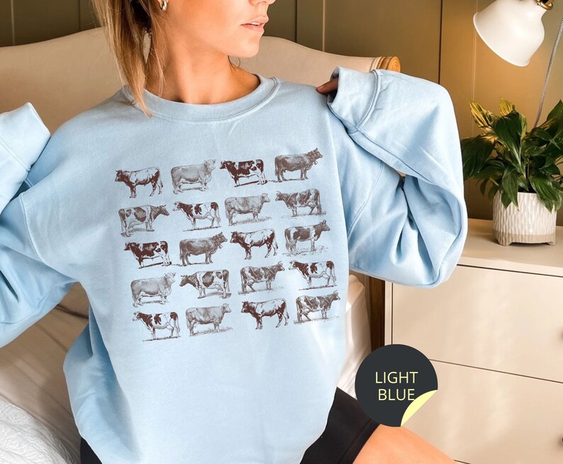 May include: A light blue sweatshirt with a pattern of brown cows printed on the front. The sweatshirt has a crew neck and long sleeves.