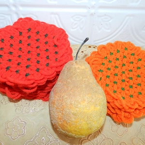May include: A still life featuring a decorative pear and two crocheted coasters. One stack of coasters is bright red, the other is orange. Both have green accents. The pear is yellow and covered in glitter.