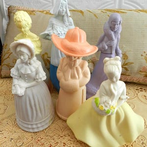 May include: A selection of vintage figurines of women in period clothing. The figurines are in pastel shades of white, yellow, peach, and lilac. Some figures are wearing bonnets, hats, and long dresses, evoking a bygone era.