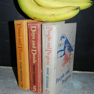 May include: Three vintage books with titles "Times and Places", "Days and Deeds", and "People and Progress" are stacked together. The books are in shades of orange, red, and off-white. Two bananas rest on top of the books. The books are part of the "Basic Readers" series.