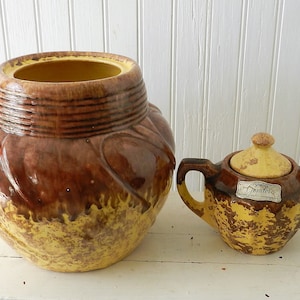 May include: A set of two vintage ceramic cookie jars with a brown and yellow glaze. The larger jar has a rounded shape with a wide opening. The smaller jar has a lid and two handles. Both jars have a label that reads "Gourde's".