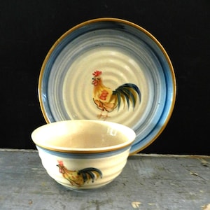 May include: A ceramic bowl and plate set with a rooster design. The plate has a blue and white spiral pattern with a gold rim. The bowl and plate feature a colorful rooster illustration. The bowl has a blue stripe around the rim.