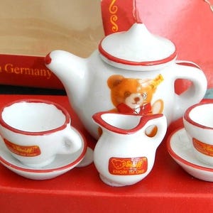 May include: A miniature white porcelain tea set with red trim, featuring a teapot, creamer, and two teacups with saucers. The teapot has a teddy bear design. The set is displayed on a red box, with the text "handbemalt - made in Germany".