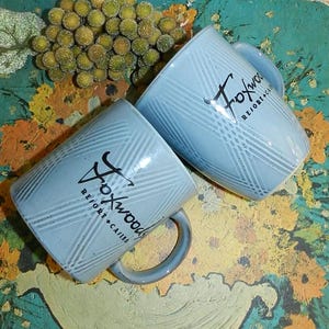 May include: Two light blue ceramic mugs with the text "Foxwood Resort + Casino" in black. The mugs have a plaid pattern and a curved handle. The mugs are on a colorful, patterned surface.