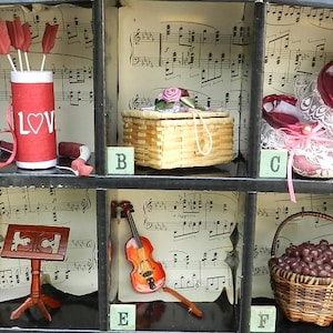 Puede incluir: Una vitrina con seis compartimentos, cada uno con un objeto decorativo diferente. Los artículos incluyen un cilindro rojo con "LOV", una cesta tejida, zapatillas de ballet rojas, un atril, un violín y una cesta de esferas marrones.