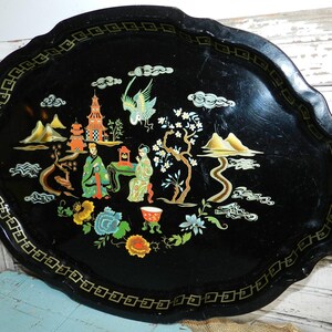 May include: A black oval serving tray with a decorative Asian-inspired design. The tray features a detailed landscape scene with figures, pagodas, and floral elements in various colors. The tray's edge has a gold geometric border.