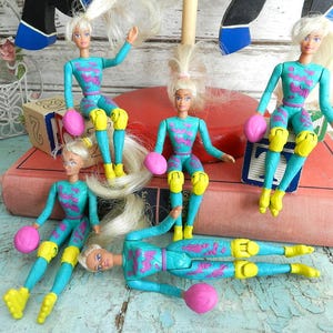 Op de afbeelding: Een verzameling vintage rolschaatspoppen. Elke pop draagt een turquoise en paars jumpsuit met gele kniebeschermers en rolschaatsen. Ze hebben blond haar en roze beschermende kleding.