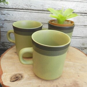 May include: Three green ceramic mugs with dark brown bands near the rim and a matching planter with a green plant. The mugs are on a wooden surface, creating a rustic aesthetic. The mugs have a handle.