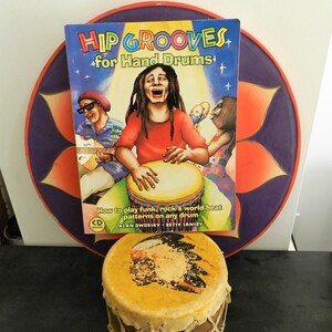 May include: A book titled "Hip Grooves for Hand Drums" with a colorful cover featuring three people playing drums. The book includes instructions on how to play funk, rock, and world-beat patterns on any drum. The book is sitting on top of a purple and orange mandala-like design.