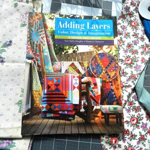 May include: A book titled "Adding Layers: Color, Design & Imagination" with 15 original quilt projects by Kathy Doughty of Material Obsession. The cover features a colourful quilt with a variety of patterns and colours.