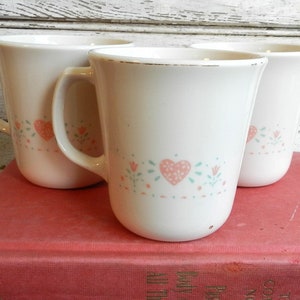 May include: Three white ceramic mugs with a pink and green floral design featuring hearts. The mugs are sitting on a red book.
