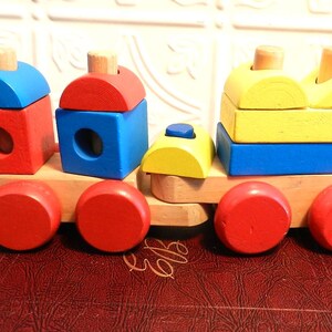 Puede incluir: Un tren de juguete de madera con bloques rojos, azules y amarillos. El tren tiene cuatro ruedas y una pila de bloques en cada vagón. Los bloques tienen forma de cuadrados y círculos.