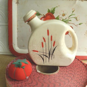May include: Cream-colored ceramic pitcher with a handle and a cork stopper. The pitcher is decorated with a design of red cattails and black stems. A red tomato pin cushion and a roll of red ribbon are in the foreground.