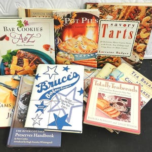May include: A collection of cookbooks with titles like "Bar Cookies A to Z", "Pot Pies", "Savory Tarts", and "Bruce's Bakery Cookbook". The books are stacked on a dark surface, showcasing various recipes and culinary themes.