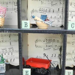 Puede incluir: Una caja decorativa con objetos en miniatura. Incluye una cesta, un cuenco con un rodillo, una máquina de coser, un carro rojo, una máquina de chicles y una botella de vidrio. El fondo presenta partituras musicales.