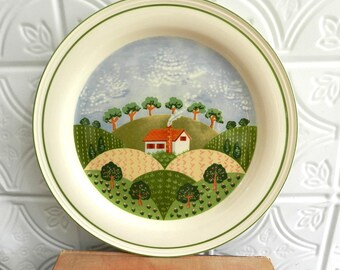 May include: A round decorative plate with a painted landscape scene. The scene features a white house with a red roof, green hills, and trees under a cloudy blue sky. The plate has a cream-colored border with a green trim.