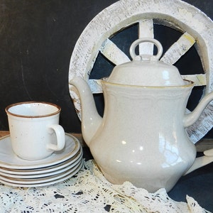 May include: A beige teapot with a brown trim and a matching cup and saucer. The teapot has a lid with a knob on top. The cup and saucer are stacked on top of each other. The teapot and cup are on a white lace tablecloth.