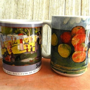 May include: Two ceramic coffee mugs. One mug features a colorful painting of people in a park, with the text "The Art Institute of Chicago". The other mug has a painting of red and yellow fruit.