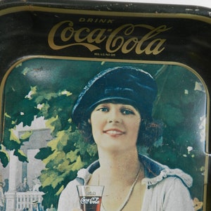 May include: Vintage Coca-Cola metal tray featuring a woman in a blue hat holding a glass of Coca-Cola. The tray has a green background with a leafy design and the text "Drink Coca-Cola" in gold lettering.