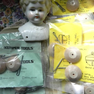 May include: A close-up of a doll head with a porcelain face and a white dress. The doll head is surrounded by packaging for doll making supplies, including wooden buttons, pins, and instructions for assembling doll legs. The packaging includes the text "Kemper Tools" and "Bend Legs".