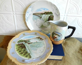 May include: Three vintage ceramic plates with images of Niagara Falls. The plates are white with blue and gold trim. The plates are arranged on a brown burlap surface with a blue book underneath.