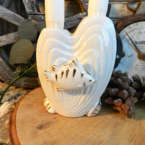 Puede incluir: Candelero de cerámica blanca en forma de corazón con dos ranuras para velas y un diseño de pez pintado en oro. El candelero está sobre una superficie de madera.