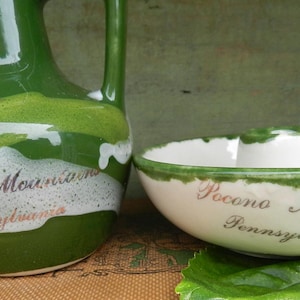 May include: A green and white ceramic pitcher and a matching bowl, both featuring the text Pocono Mountains Pennsylvania in a script font. The pitcher has a handle and a narrow neck, while the bowl has a wide rim with green painted accents.