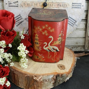 May include: Red metal tin with a gold painted design of a pagoda and two flamingos. The tin has a rounded top and a rectangular base. The tin is sitting on a wooden slice.