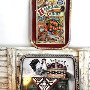 Puede incluir: Dos bandejas rectangulares decorativas. La bandeja superior tiene un borde burdeos y presenta el texto "The Heartland Collection" con una ilustración de cerezas. La bandeja inferior tiene un gallo y una cesta de manzanas.