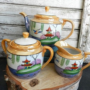 Puede incluir: Un juego de té japonés vintage con un esmalte dorado y blanco. La tetera, el azucarero y la jarra de crema presentan escenas pintadas a mano de pagodas, flores de cerezo y pájaros.