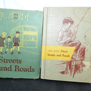 Puede incluir: Dos libros infantiles antiguos. El libro de la izquierda se titula "Streets and Roads" con ilustraciones de niños y un autobús. El libro de la derecha se titula "The New More Streets and Roads" con un niño pescando. Ambos libros están desgastados.