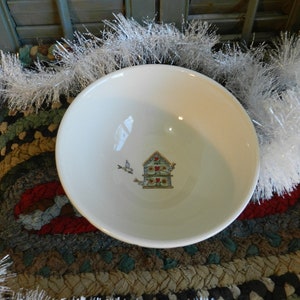 May include: White ceramic bowl featuring a blue and red birdhouse design. The birdhouse has a red roof and white trim. Two small birds are depicted flying around the birdhouse.