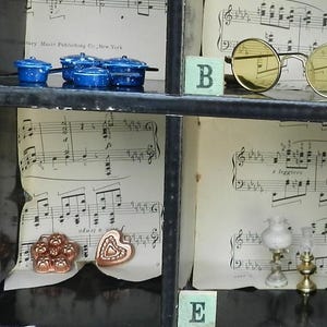 Puede incluir: Una vitrina con objetos en miniatura. Estante superior: ollas azules con lunares blancos y gafas redondas de color amarillo. Estante central: objetos en forma de flor y corazón de color cobre. Estante inferior: dos lámparas en miniatura. Fondo: partituras.