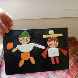 May include: A wooden toy puzzle with two figures made of geometric shapes. The figures are a boy with a green hat and a girl with a yellow hat. The puzzle pieces are on a black background.