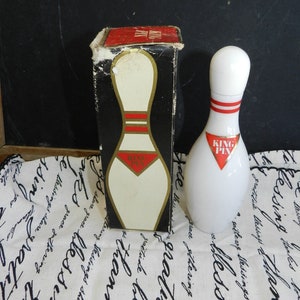 Peut inclure: Une bouteille en céramique blanche en forme de quille de bowling avec un motif de rayures rouges et blanches et les mots "KING PIN" en rouge sur une étiquette en forme de losange. La bouteille est posée sur un tissu blanc avec des inscriptions noires. La bouteille est à côté d'une boîte avec un dessus rouge et une quille de bowling blanche sur le devant.