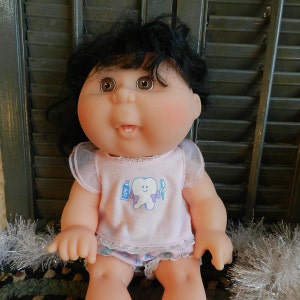 Peut inclure: Une poupée Cabbage Patch Kid aux cheveux noirs et aux yeux bruns portant une chemise rose avec un motif de dent et le texte "CPK" dessus. La poupée est assise sur une surface blanche et moelleuse.