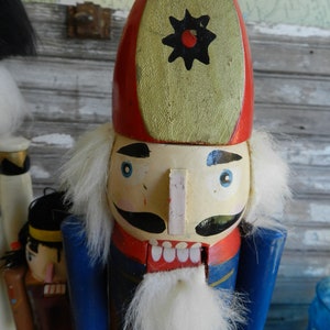 May include: A wooden nutcracker with a red and white hat, blue jacket, and white beard. The nutcracker has a painted face with blue eyes and a black mustache. The hat has a black star with a red center.