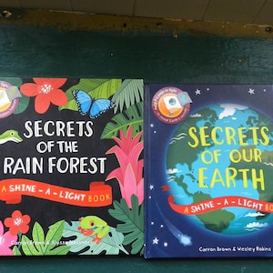 May include: Two children's books titled "Secrets of the Rain Forest" and "Secrets of Our Earth." The first book features a black cover with colorful illustrations of rainforest flora and fauna. The second book has a blue cover with a globe illustration.